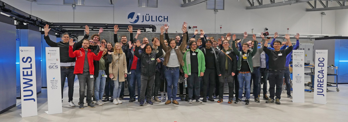 Group of Hackathon participants jumping in front of JUWELS Cluster in the machine hall